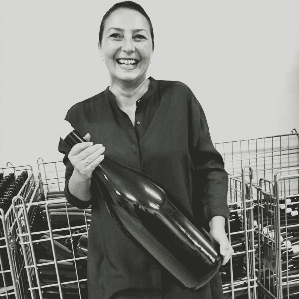 The founder of VinoRoma, Hande Leimer, who is holding an oversized bottle of wine during a visit with members to the cellar of a natural winery.