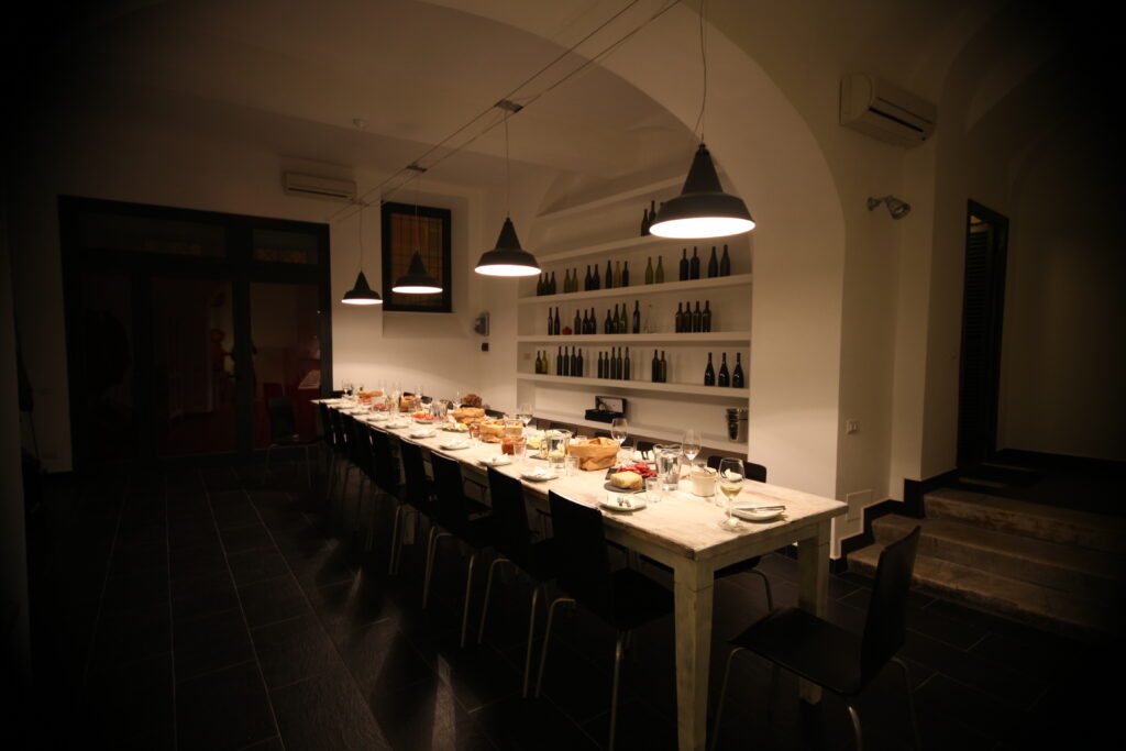 Sala di degustazione di VinoRoma con luce soffusa e il tavolo di degustazione apparecchiato con salumi, formaggi, pane, piatti e bicchieri di vino.