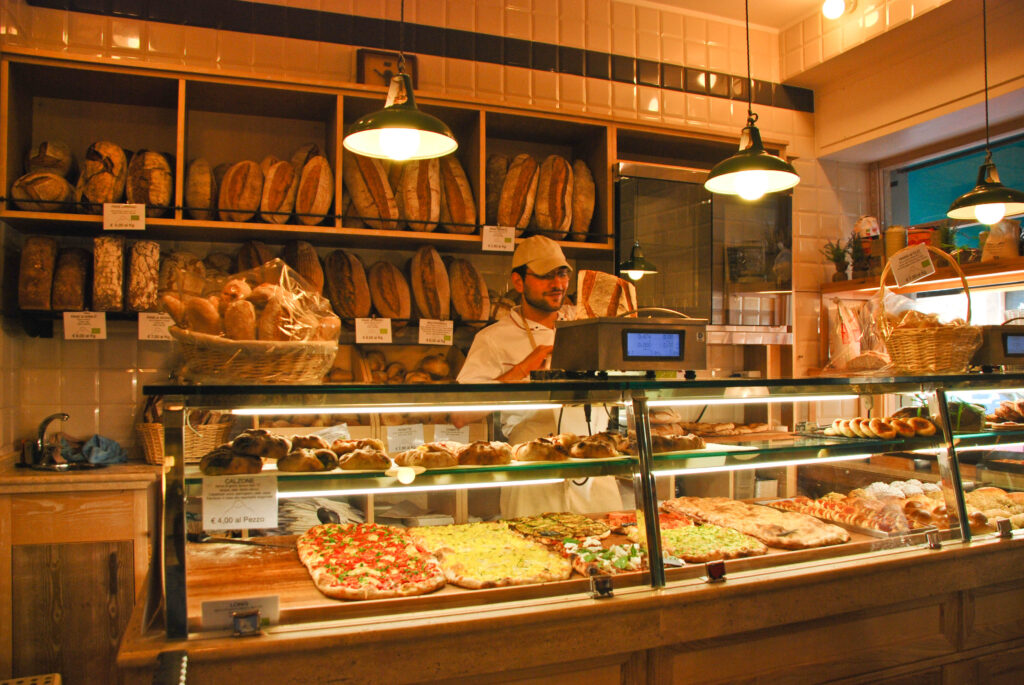 A baker weighing bread behind a counter filled with pizza a soft lighting in a bakery during a walking food tour with members.